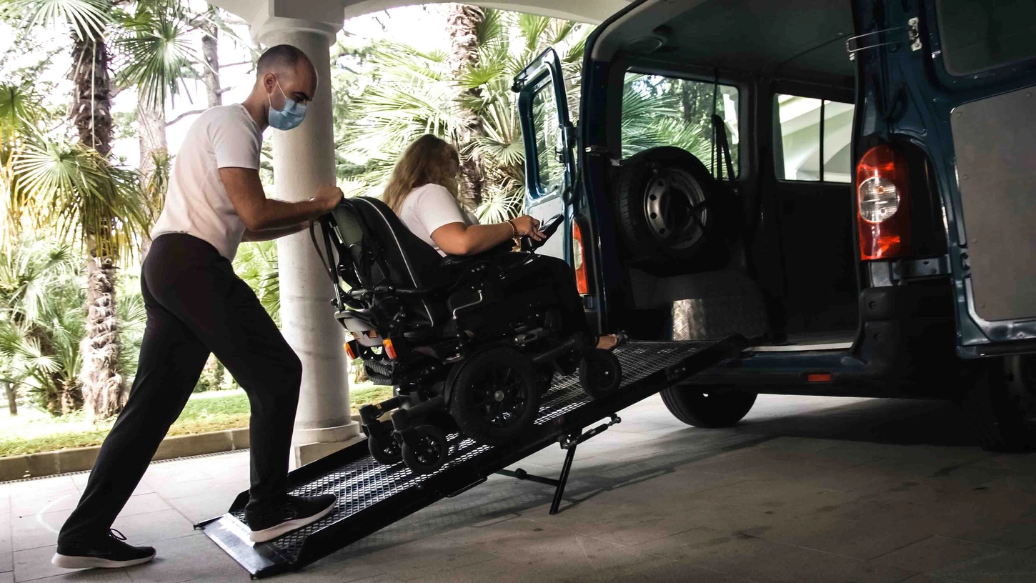 Handicap Fahrzeuge GmbH - Unterstützung einer behinderten Frau im Rollstuhl beim Transport mit einem barrierefreien Fahrzeug mit Rampe, die eine Gesichtsmaske trägt.